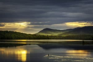 Sunrise over distant mountains reflected in glassy lake with a single loon swimming